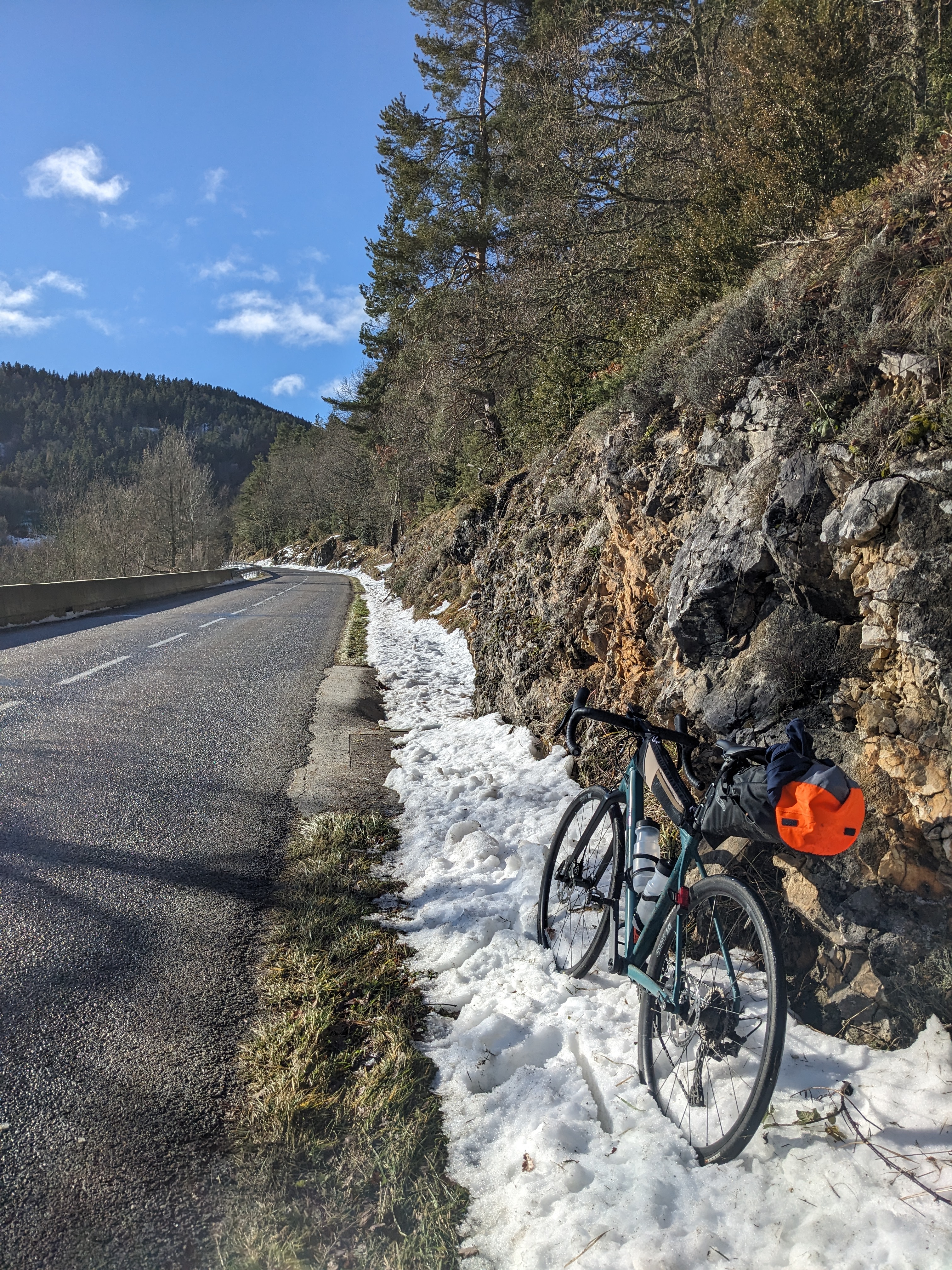 A bicycle rests on a patch of snow by the roadside, adjacent to a rocky and forested hillside. The road curves gently and is bordered by grass and patches of snow. Tall evergreen trees line the right side, with a clear blue sky overhead. A bright orange item is strapped to a saddle bag, contrasting with the natural surroundings.